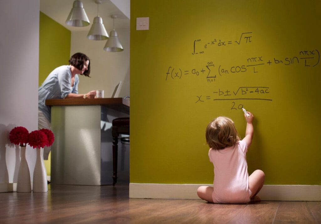 A toddler sitting on the floor works out complex mathematical equations on the wall while her mother nearby is oblivious